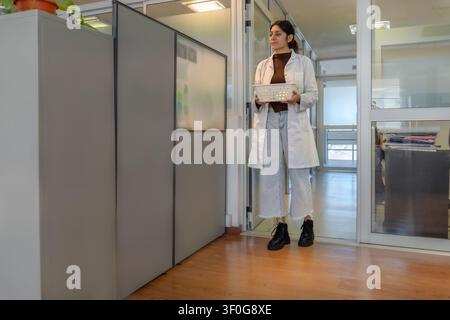 Young female scientist in white lab coat standing in a glass doorway of a bright institutional office, holding an empty white plastic tray with cubicl Stock Photo