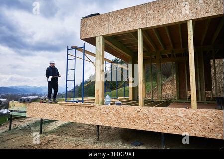 Foreman examines design documents on elevated wooden platform, coordinating house construction and monitoring progress in countryside setting Stock Photo