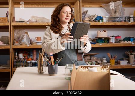 Pottery Studio Owner Using Tablet in Ceramic Workshop Surrounded by Shelves and Tools Stock Photo
