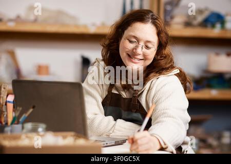 Smiling Female Potter Working in Her Pottery Studio Laptop and Tools While Running a Small Business Stock Photo
