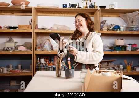 Smiling Female Ceramic Artist Using Tablet in Pottery Studio As Small Business Owner Managing Orders Stock Photo