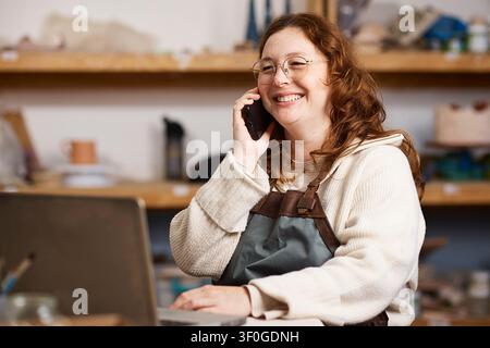 Smiling Pottery Studio Owner Talking on Phone While Working on Small Business at Laptop Stock Photo