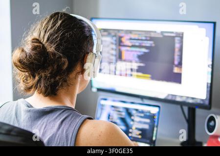 Man wearing wireless headphones works at a desk with laptop and external monitor in a home office. Concept of remote work, software development, produ Stock Photo