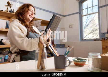 Smiling Pottery Studio Owner Using Tablet in Bright Ceramics Workshop - Small Business Creative Artist Stock Photo