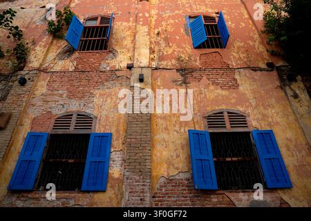 A low-angle exterior shot of a weathered vintage building wall, featuring exposed red brickwork under cracked stucco plaster, open bright blue wooden Stock Photo