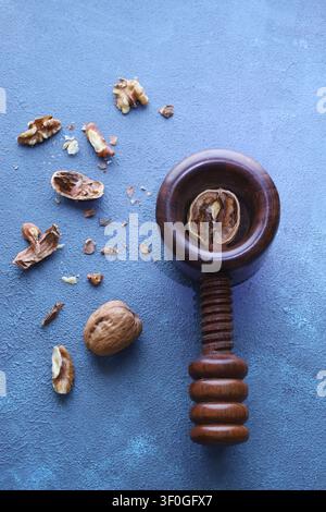 Top view of a dark handcrafted wooden nutcracker surrounded by whole walnuts and broken shells on a blue background. Stock Photo