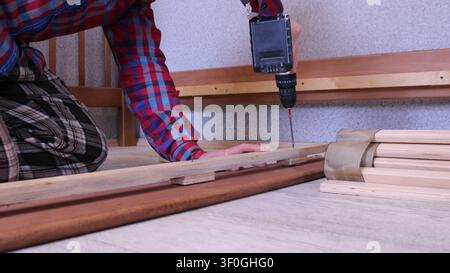 man assembles a bed frame, driving a screw into a wooden slat using a cordless drill. A finished block of wooden slats, bound tightly together Stock Photo