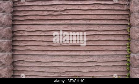 brown fence made of concrete sections imitating wood planks with a textured surface. Stone pillars are visible on the sides, and a green ivy branch Stock Photo