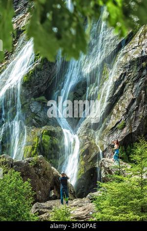 Man photographing woman posing on rocks in front of powerful Powerscourt  waterfall cascade in Ireland forest. Stock Photo