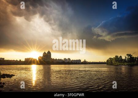 Dramatic sunset after a thunderstorm over Obolon Embankment in Kyiv, Ukraine. Golden sunbeams break through storm clouds, reflecting across the Dnipro Stock Photo