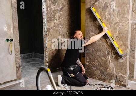 Builder checking the evenness of marble tiles with a level tool, ensuring precision and accuracy during renovation work on a wall Stock Photo