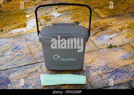 Small grey indoor plastic caddy with lockable lid for domestic food waste plus a roll of disposable plastic bags on a wet garden patio background Stock Photo