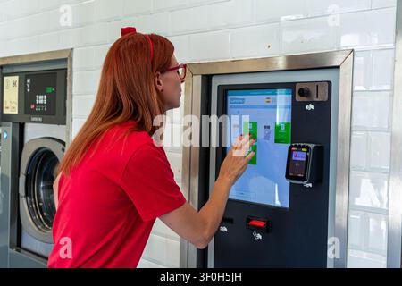 Woman with red hair and glasses choosing a washing program on the touchscreen of a modern self service laundromat machine, simplifying laundry tasks a Stock Photo