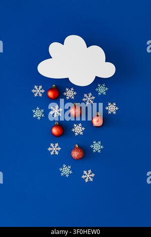 Creative winter cloud with falling snowflakes and red Christmas ornaments on blue background. Minimal Christmas and New year concept. Stock Photo