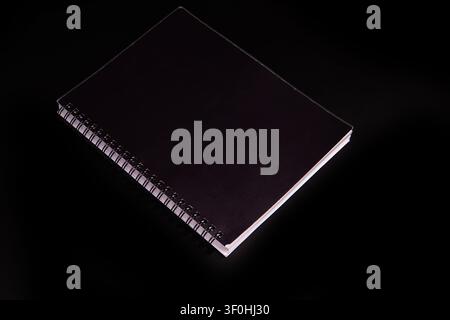 Professional spiral notebook with matte black cover, positioned diagonally on stark black background, emphasizing texture and depth, clean minimalist Stock Photo