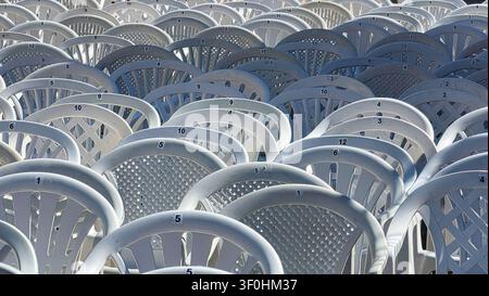 Multiple white plastic chairs tightly arranged in rows, each labeled with black numbers on small white stickers, creating a pattern of repetition and Stock Photo
