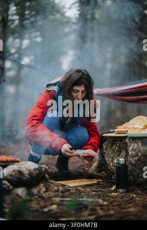 Woman preparing food by a campfire in a forest campsite wearing a red jacket and scarf Stock Photo