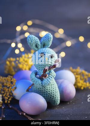 Cute knitted stuffed Easter bunny toy and painted eggs, close-up, bokeh colored background with garland Stock Photo