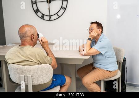 A senior couple sits at a table in a bright room. They drink tea or coffee and talk. The man holds a cup while the woman listens. A clock is on the wall behind them. Stock Photo