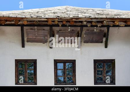 View of a traditional house facade with a stone roof, wooden support beams, and three dark-framed windows against a white plastered wall in Bosnia and Stock Photo