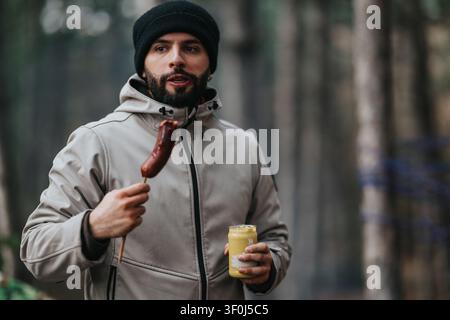 Bearded man eating a sausage with mustard in a forest while wearing a hooded jacket and beanie Stock Photo
