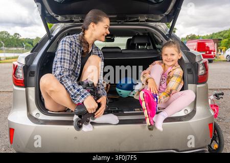 Mother and daughter putting on inline skates while sitting in the open car trunk before skating together. Active family lifestyle and quality time Stock Photo