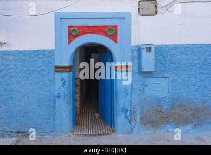 View of a traditional blue-painted doorway with a red and green arched frame set into a textured wall in Taghazout, Souss-Massa, Morocco. Stock Photo