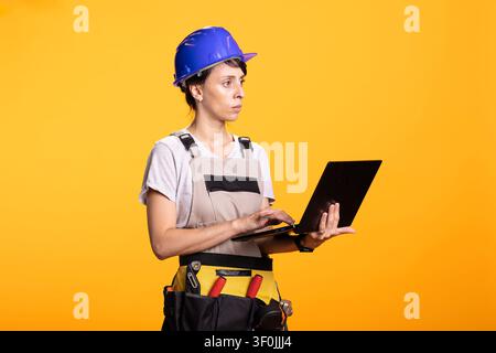 Caucasian female builder holding laptop reviewing job information for repair work. Portrait of construction worker against yellow background preparing site documentation and workflow planning. Stock Photo