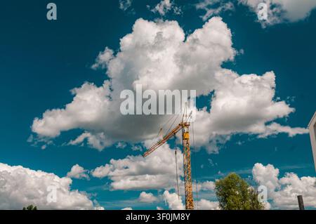 A tall yellow tower crane stands against a bright blue sky filled with large, fluffy white cumulus clouds. The scene is set outdoors during the day. Stock Photo