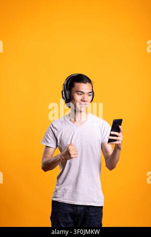 Asian man wears wireless headphones while answering video call on smartphone. Focused male individual smiles and communicates online, engaging in conversation through modern digital technology. Stock Photo