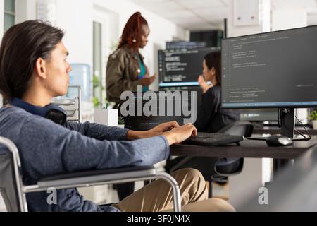 Asian man in wheelchair reviews frontend and backend code on monitor at desk. Confident IT professional symbolizes inclusive workplaces, innovation and problem solving in tech. Stock Photo