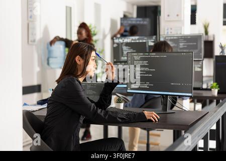 Asian software developer reviewing coding scripts on desktop monitor with focused, thoughtful expression. Digital workspace represents debugging, analytics and strategic problem solving in tech startup workspace. Stock Photo