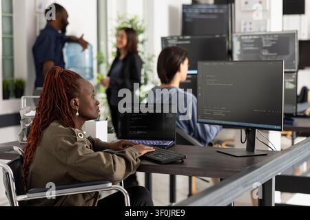 Black female programmer in wheelchair coding at desktop pc in inclusive workplace. Diverse tech team working in professional software development environment with multiple monitors. Stock Photo