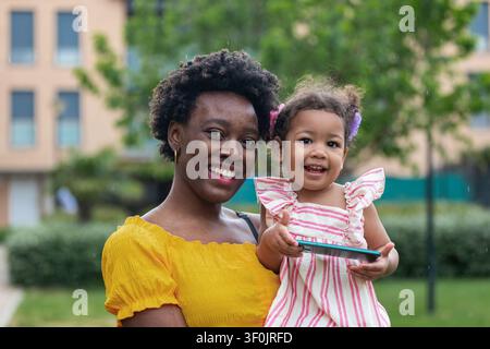 Cheerful mum holding her toddler daughter in a city park, both smiling at the camera. Family portrait with greenery and urban buildings in soft focus, Stock Photo