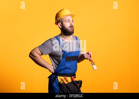 Construction worker stands with paintbrush, appearing deep in thoughts about renovation decisions. Caucasian male builder reflecting on painting work tasks against yellow studio background. Stock Photo