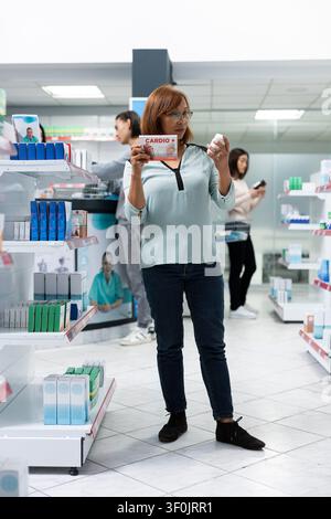 Asian man stands in local pharmacy holding prescription medication and supplements. Focused male customer reviews product labels, representing medical retail, treatment choices and lifestyle health. Stock Photo