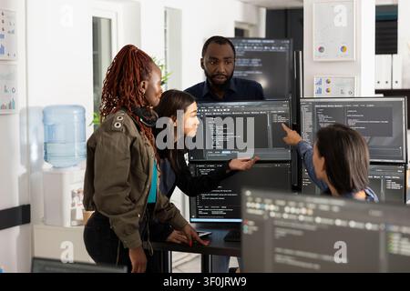Diverse group of software developers in modern office collaborating, discussing and analyzing source code on multiple computer monitors, working together on web development project. Stock Photo