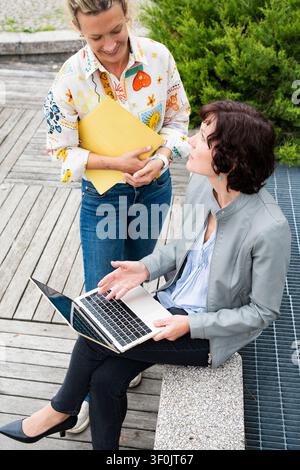 A senior businesswoman actively engages with a colleague outdoors, discussing strategy while working on a laptop. They are casually dressed and appear Stock Photo