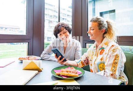 Two women enjoy a lunchtime break at a modern cafe, sharing a casual conversation over a meal with pizza. Their business attire suggests a professiona Stock Photo