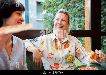 Two women take a break from work, sharing laughter and pizza at a cozy cafe. One holds a slice, smiling widely, capturing the moment with joy and cama Stock Photo