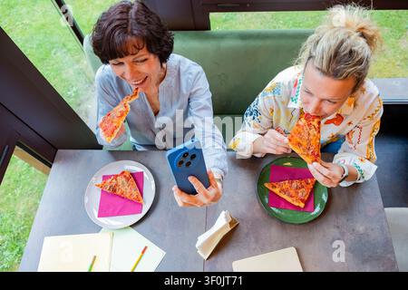 Two businesswomen sit together at a cafe table, enjoying pizza slices during a lunch break. One woman takes a selfie with her phone, capturing the liv Stock Photo