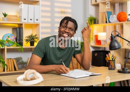African American young man on home table writes notes and plans ideas with pen during focused work. Black guy on home table develops study plan in notebook and shapes goals for remote job or course. Stock Photo