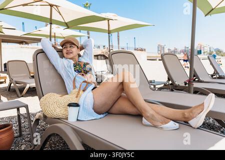 Mature woman wearing a cap, lounging on a sunbed at a beach, enjoying a sunny day. Umbrellas provide shade, while she relaxes with a straw bag and bev Stock Photo