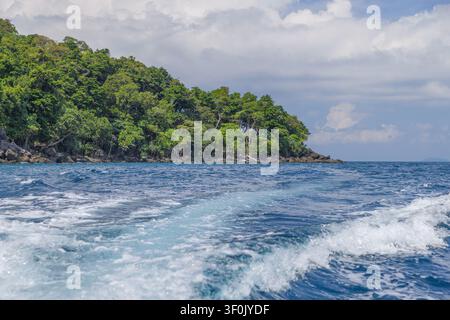 Lush green tropical forest lines a rocky shoreline on Pulau Weh, Indonesia, viewed from the open sea with foamy boat wake, deep blue water, and cloudy Stock Photo