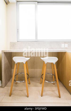 Bright minimal kitchen corner with stone counter top breakfast bar, two white modern stools with wooden legs, tiled floor, and large window. Stock Photo