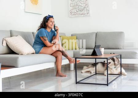 A cheerful woman talking on her phone while working from home, seated on a cozy sofa with a laptop. Her pet dog relaxes by the coffee table, creating Stock Photo