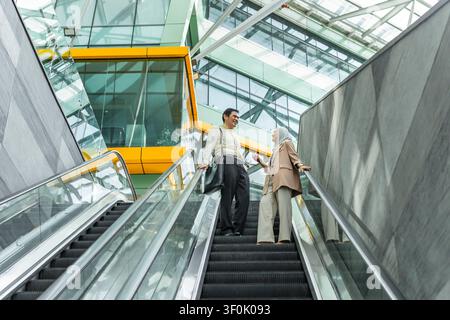 Two Asian business colleagues talk while riding an escalator in a bright modern glass atrium, with sleek architecture, reflections, and an urban offic Stock Photo