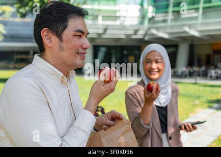 Two Asian coworkers chat and smile while eating apples during a relaxed lunch break in an office courtyard, holding a paper bag lunch, in casual busin Stock Photo