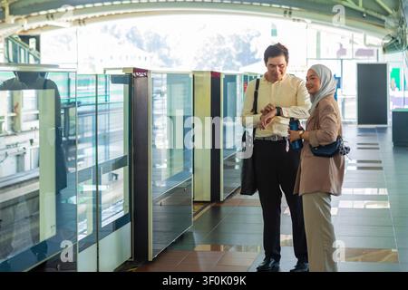 Two office colleagues waiting, checking the time and chatting while holding a bottle and bag. Modern setting with glass barriers and bright light. Stock Photo