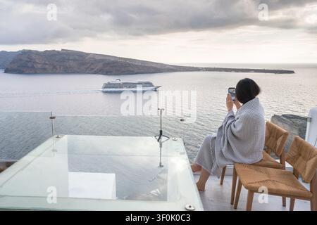 Woman in a robe sits on a luxury Santorini hotel terrace, photographing a cruise ship on the calm Aegean Sea at sunset, with cliffs and cloudy sky. Stock Photo